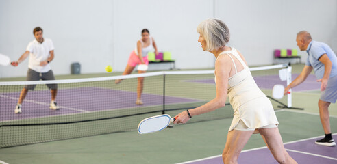 Focused active aged woman playing friendly pickleball match on indoor court, rear view. Senior...