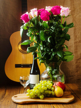 Still life with grapes, wine bottle and glasses, pear, bouquet of roses and classic wooden guitar on wooden table. Vertical image.
