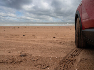 Side of a red car parked on a sandy beach with stunning view on the ocean. Travel and tourism concept. Renting vehicle for a trip theme. Cloudy sky and water surface in the background.