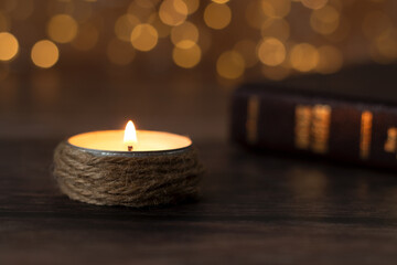 Candle burning with bright flame and closed holy bible book on wooden table with bokeh background. Close-up. Selective focus. Spiritual light shining in the darkness, Christian biblical concept.