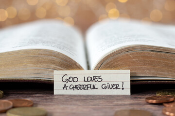 God loves a cheerful giver, handwritten text note in front of open holy bible book and golden coin...