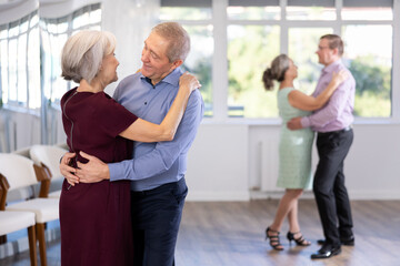 Elderly married couple is leisurely dancing slow dance in studio for beginners learning pair dancing. Woman clung to her partner, put her head on mans shoulder