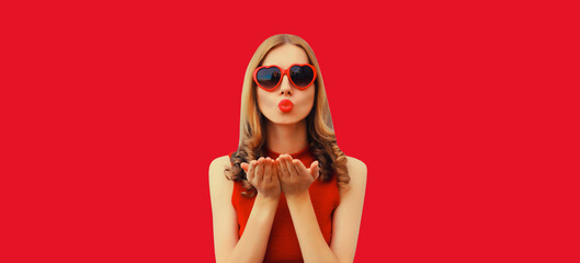 Portrait of beautiful young woman blowing her lips with red lipstick sends sweet air kiss wearing heart shaped sunglasses on studio background - obrazy, fototapety, plakaty