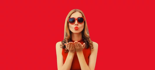 Fotobehang Portrait of beautiful young woman blowing her lips with red lipstick sends sweet air kiss wearing heart shaped sunglasses on studio background © rohappy