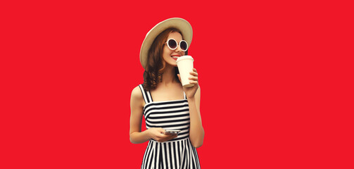 Portrait of happy smiling young woman drinking fresh juice or coffee and looking away wearing summer straw hat, striped dress on red studio background