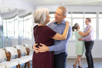 Obraz premium Elderly married couple is leisurely dancing slow dance in studio for beginners learning pair dancing. Woman clung to her partner, put her head on mans shoulder