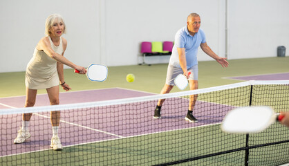 Portrait of sporty senior woman playing doubles pickleball with male partner on indoor court, ready to hit ball. Sport and active lifestyle concept