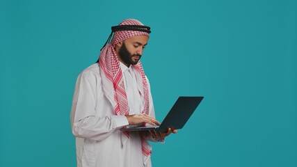 Model using pc touchpad in studio, dressed in traditional islamic costume and kufiyah. Middle eastern person holding modern sleek laptop and scrolling websites or communicating online.
