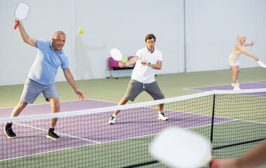 Obraz premium Portrait of sporty senior man playing doubles pickleball with male partner on indoor court, ready to hit ball. Sport and active lifestyle concept..