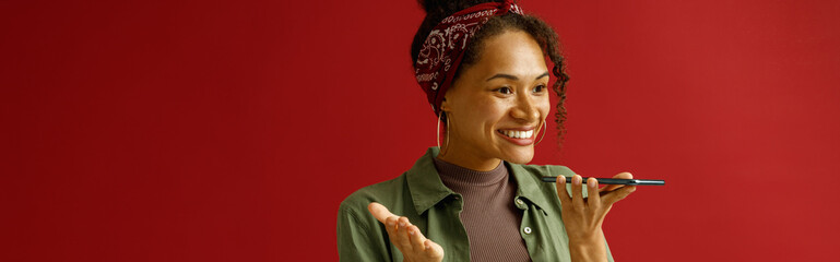 Positive woman is recording audio message on mobile phone while standing on red studio background