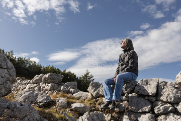 Adult gray-haired man sitting on a rocky fence against a cloudy sky and looking at the clouds,...