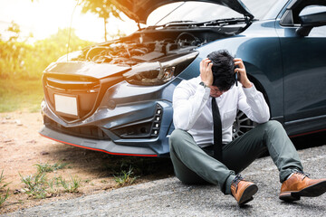 Young businessman wearing white shirt and call roadside assistance. Worried mature man holding his head by hands sitting near his broken car with raised hood on the roadside of a rural road.