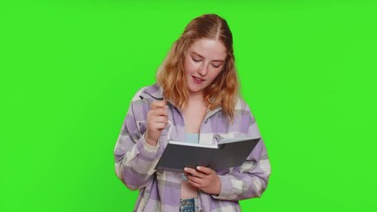 Thoughtful journalist young woman making notes, writing down thoughts with pen into notepad notebook diary, to do list, good idea. Pretty redhead student girl isolated on green chroma key background