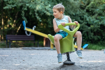 Naklejka premium A little boy sits on a green rocking chair swing in the playground and looks away