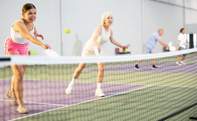 Expressive energetic girl playing doubles pickleball match in team with older woman indoors,...