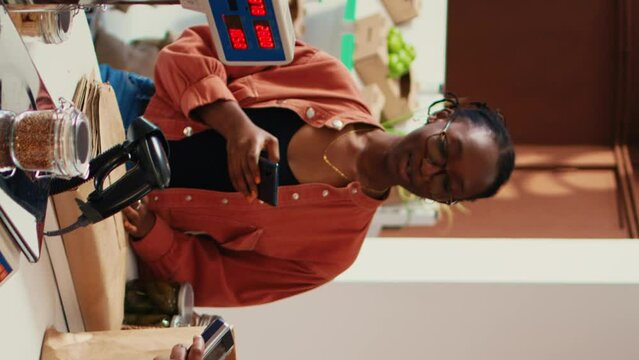 Vertical Video Client using nfc payment at pos to buy eco products, visiting local farmers market to restock pantry essentials. Vegan woman paying with mobile phone at checkout, nonpolluting goods