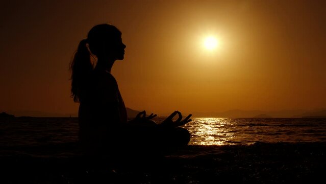 Girl silhouette doing tranquil namaste. A calm teen silhouette training yoga and enjoy the warm sea evening during summer weekend.