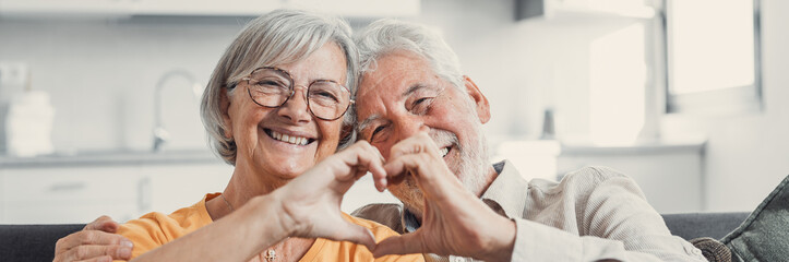 Close up portrait happy sincere middle aged elderly retired family couple making heart gesture with fingers, showing love or demonstrating sincere feelings together indoors, looking at camera..