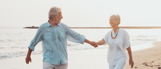 Couple of old mature people walking on the sand together and having fun on the sand of the beach enjoying and living the moment. Two cute seniors in love having fun. Barefoot walking on the water. - obrazy, fototapety, plakaty