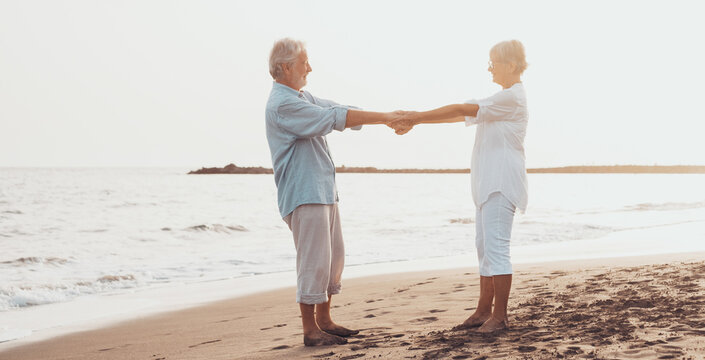 Couple of old mature people dancing together and having fun on the sand at the beach enjoying and living the moment. Portrait of seniors in love looking each others having fun.