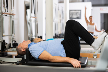 Focused mature man lying with her legs and arms suspended while doing Pilates exercise on reformer...