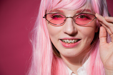 Close-up portrait of a young woman with braces in a pink wig and sunglasses on a pink background.