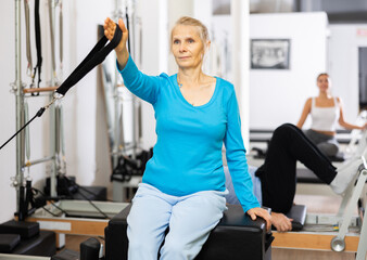 Focused elderly woman holding hand in adjustable sling during workout on Pilates reformer combo bed...
