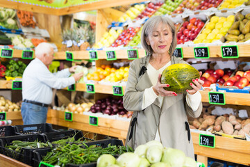 Woman choosing melon while standing in greengrocer. Old man shopping in background.