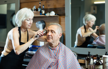 Haircut in a stylish barbershop - an elderly man sits in a chair, a woman cuts her hair and does hair styling - obrazy, fototapety, plakaty