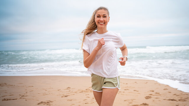Smiling female athlete jogging on sandy sea beach. Fit woman in white top and sporty shorts. Active lady running along shoreline represents healthy lifestyle. - Powered by Adobe