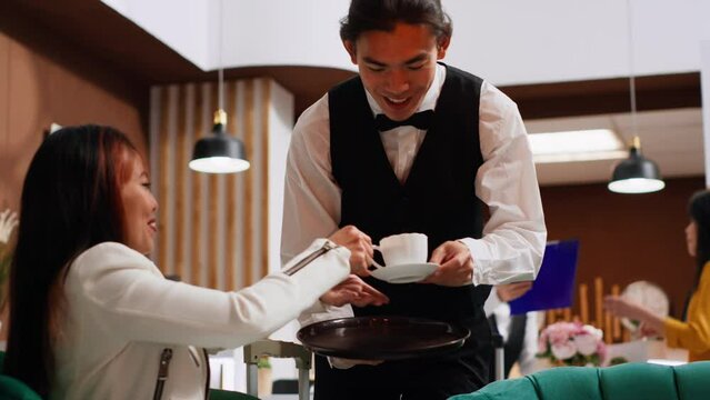 Young waiter serving coffee for asian guest in lounge area, providing excellent customer service at five star hotel. Professional concierge staff ensuring relaxed stay for traveler. Handheld shot.