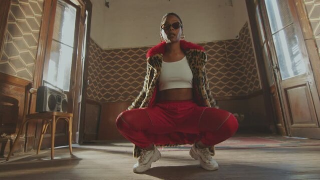 Young cool female dancer wearing sunglasses and stylish fur coat with leopard print crouching on the floor and dancing on camera in room with retro interior