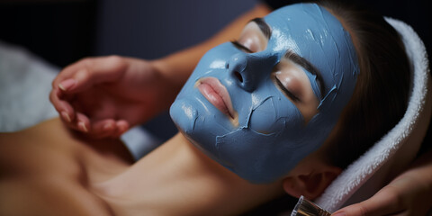 A young woman at a beauticians appointment. The specialist applies a cream mask to the face. Facial skin care and protection. A girl at the cosmetologist's office. Face in blue clay. Care procedure. B - obrazy, fototapety, plakaty