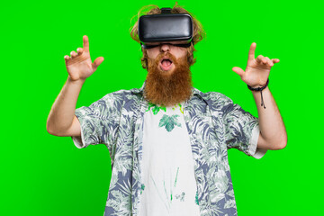 Excited happy bearded man using headset helmet app to play simulation game. Watching virtual reality 3D 360 video. Redhead guy in VR goggles isolated on green chroma key background. Future technology