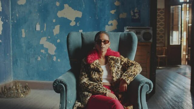 Young confident woman in fur coat with leopard print and sunglasses sitting in armchair beside retro cassette tape player and looking at camera in vintage room with cracked paint on walls