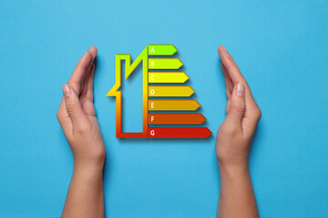 Energy efficiency rating and woman holding hands near it on light blue background, top view