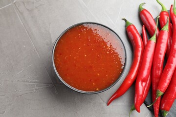 Spicy chili sauce in bowl and fresh peppers on grey table, flat lay