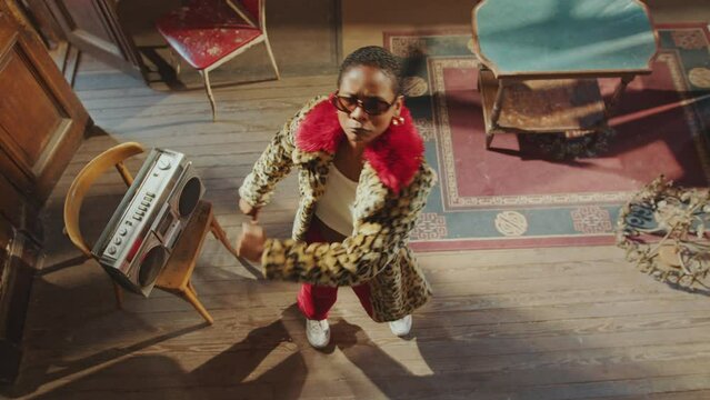 Young eccentric girl in trendy outfit looking at camera and dancing to music played by ghetto blaster in retro room. View from above