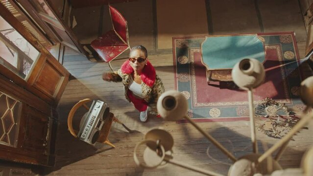 Stylish girl in trendy fur coat and sunglasses performing eccentric dance to music played by ghetto blaster in old-fashioned room. Top down view, chandelier spinning under the ceiling