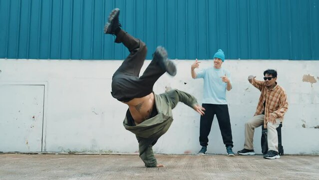 Skilled hipster doing cart wheel while multicultural friends encourage him. Hip-hop dancer moving foot step to music surrounded by group of hipster at wall near street. Outdoor sport 2024. hiphop.