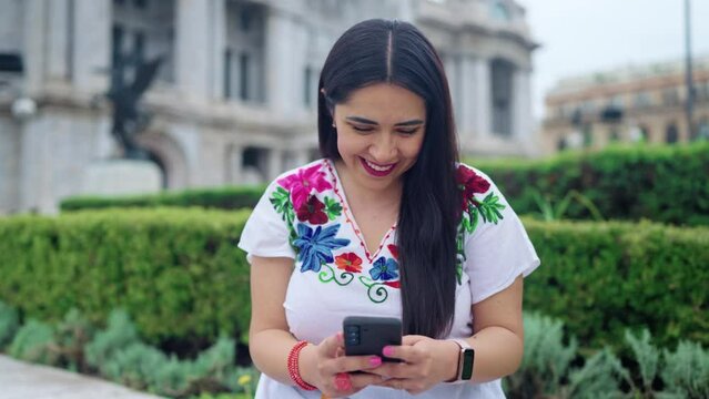 Happy young ethnic mexican woman using smartphone and looking at screen of mobile phone while sitting in front of blurred Bellas Fine Arts Palace in Mexico City and wearing traditional clothes