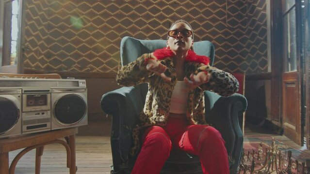 Eccentric Black girl in trendy outfit sitting in old armchair, posing on camera and performing rhythmic dance moves in sunlit retro styled room with cassette tape player. Whip zoom shot