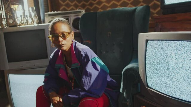Cool-looking Black girl in 80s tracksuit and sunglasses sitting among old analog TVs with static screens, posing on camera and dancing to hip-hop music