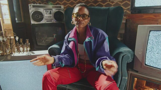 Young stylish woman in 80s outfit sitting among old TVs with static noise on screens and performing hip hop moves on camera in retro-themed studio. Whip zoom shot