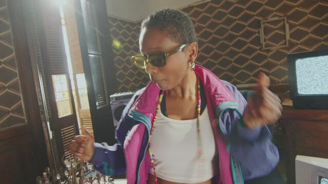 Stylish Black girl in 80s tracksuit and sunglasses standing up from armchair and dancing among analog TVs with static screens in retro-themed room. Zoom handheld shot