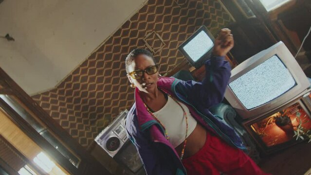 Energetic African-American girl in retro tracksuit dancing and looking at camera in old-fashioned studio with analog TVs with static screens and ghetto blaster. Shaky camera