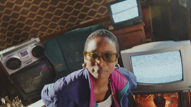 Cool African-American girl with short-haircut in 80s retro tracksuit dancing and looking at camera in old room with analog TVs with static screens in the background