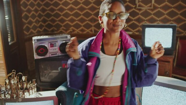 Cheerful Black girl in 80s fashion shell suit enjoying the music, dancing and smiling in room with analog TVs, cassette tape player and antique chandelier decor