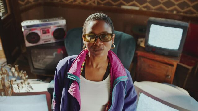 Confident Black girl in 80s outfit and sunglasses standing in retro-styled room with no signal analog TVs and posing at camera. Video portrait