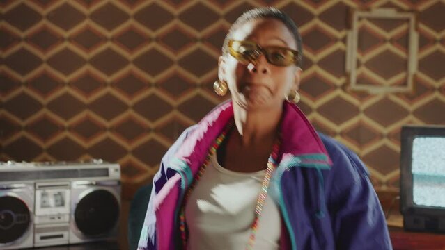 Joyous African American girl in 80s fashion shell suit and sunglasses singing with arms raised and dancing to the music in old room with analog TVs and cassette tape player
